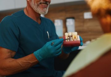 Dentist in blue scrubs showing dental implants model to patient, explaining treatment options. Office environment with professional tools in background