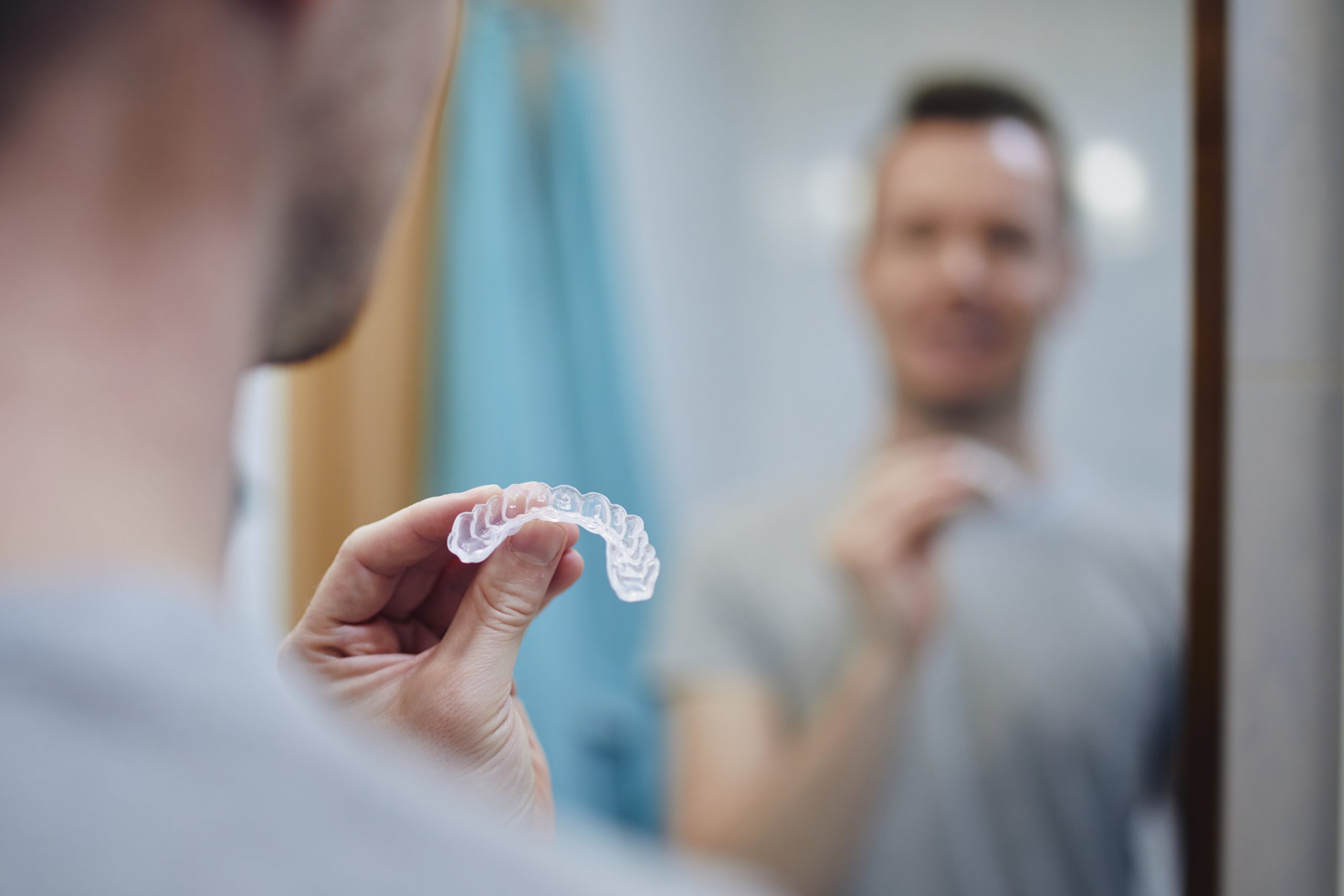 Man holding a clear custom night guard used for teeth grinding treatment in front of a bathroom mirror