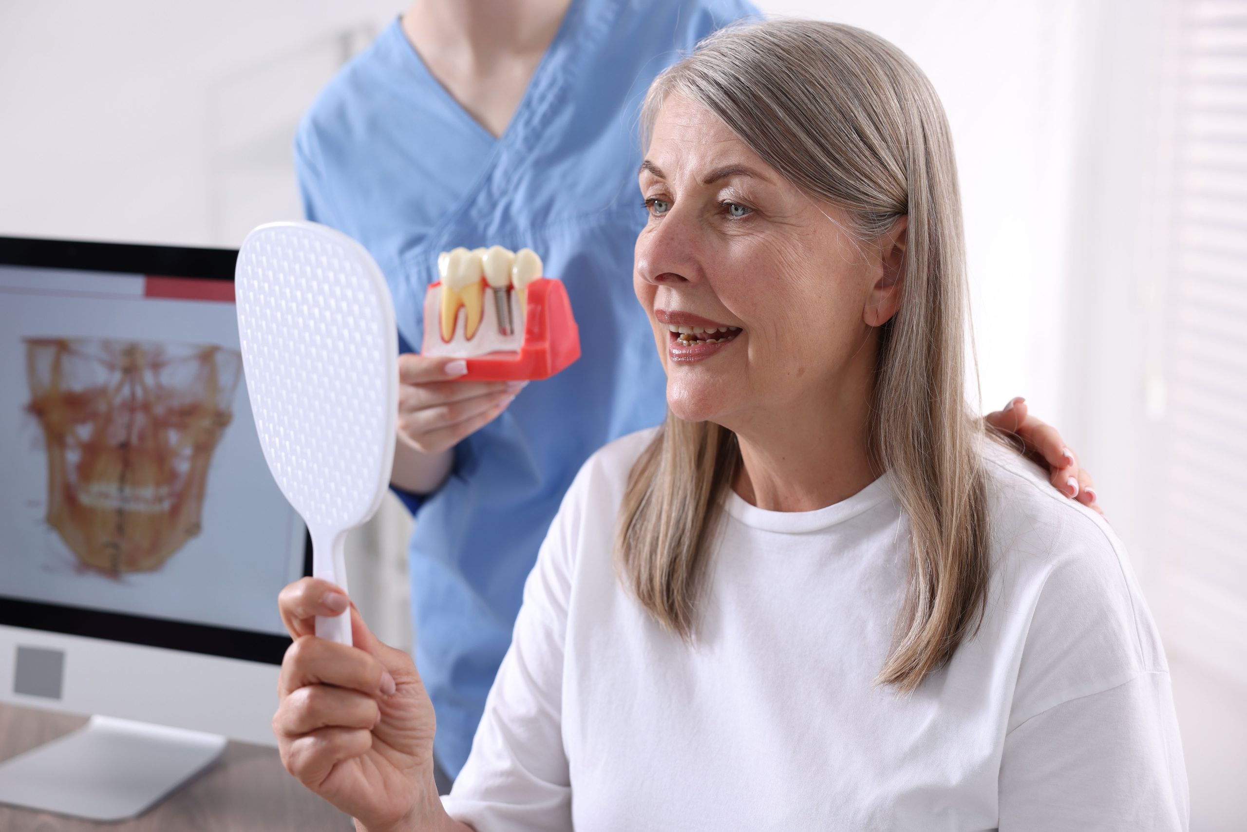 Mature woman looking in a hand mirror while a dental professional shows her a dental implants model